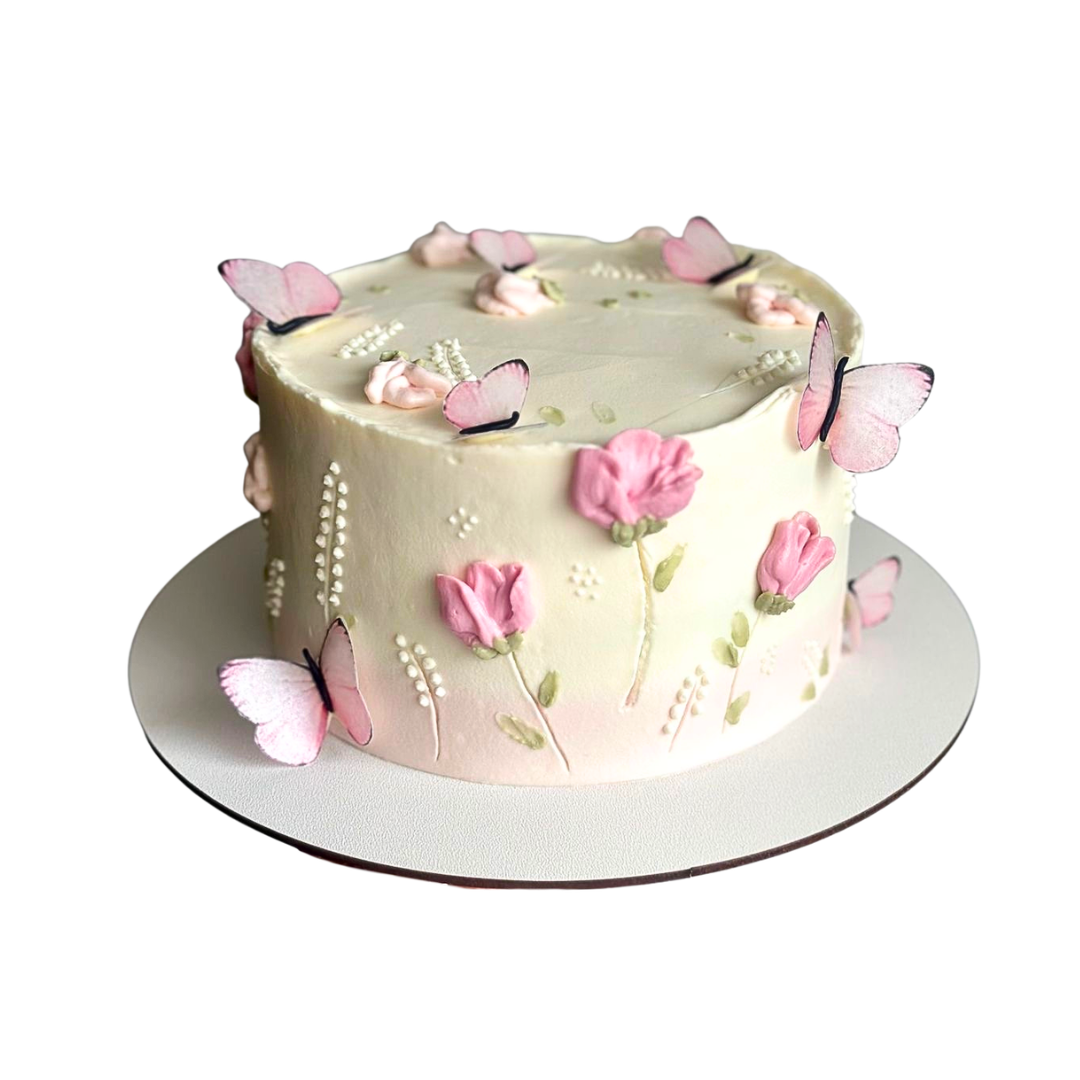 Round cream-colored cake decorated with pink wafer butterflies and piped cream flowers. Dessert Boutique.