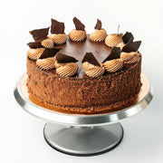 round Prague chocolate cake with piped caramel buttercream swirls, chocolate shards and crumb-coated sides on a silver cake stand against a white background