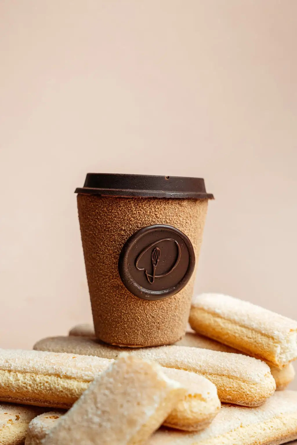 tiramisu dessert shaped like a coffee cup with cocoa-coated mousse, savoiardi biscuits and chocolate logo on top of pastries against a beige background
