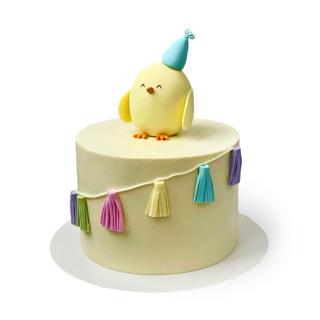 Round yellow birthday cake with a fondant chick wearing a hat and a tassel garland