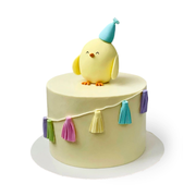 Round yellow birthday cake with a fondant chick wearing a hat and a tassel garland