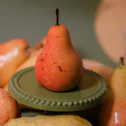 Gourmet pear-shaped dessert with chocolate stem displayed on a green mini cake stand surrounded by fresh pears