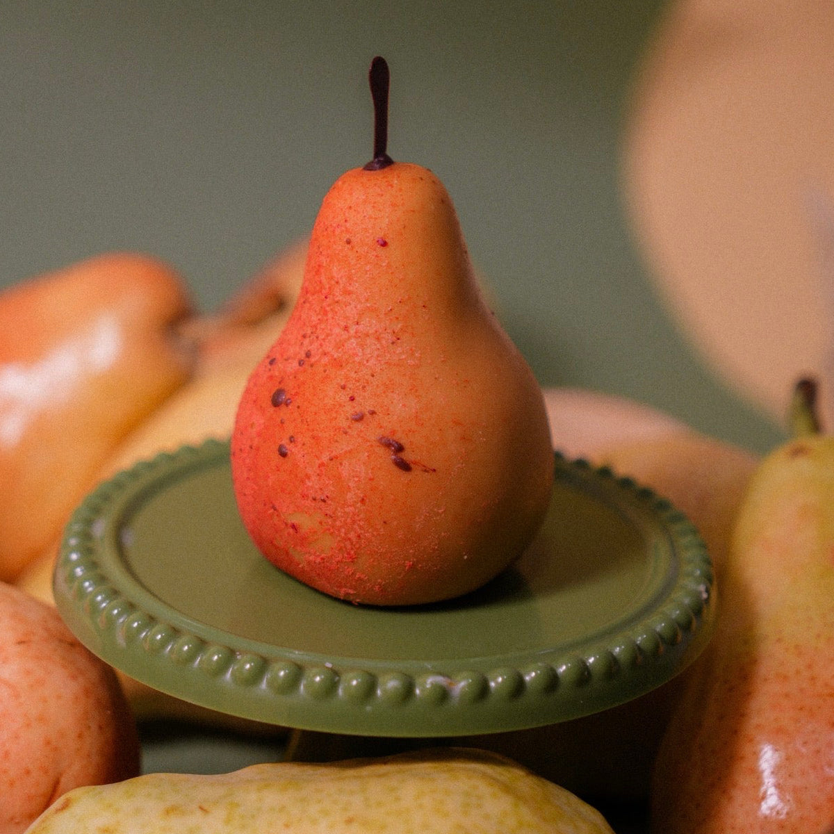 Gourmet pear-shaped dessert with chocolate stem displayed on a green mini cake stand surrounded by fresh pears