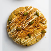 large pistachio cookie with white chocolate drizzle, roasted pistachio pieces and golden baked surface on white background