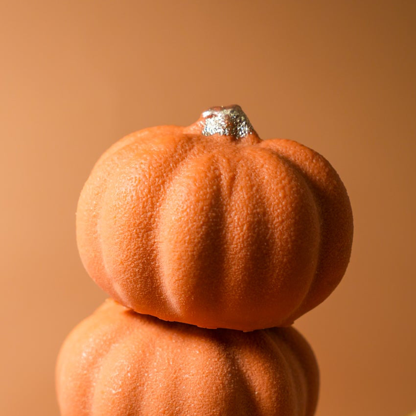 A stack of three pumpkin-shaped mousse desserts with a velvety orange finish surrounded by fresh pumpkins.⁠